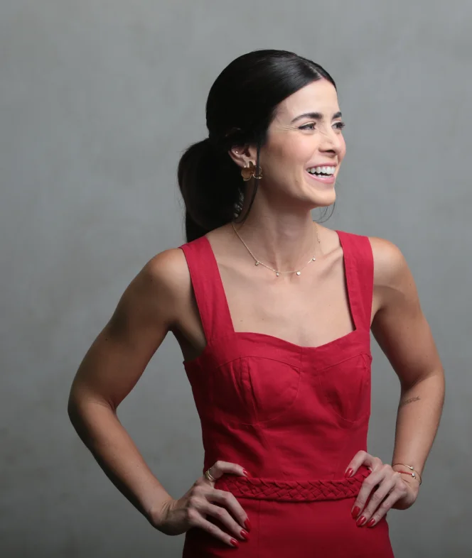 Ana Zucato, mulher branca de cabelo escuro preso, sorrindo, vestindo um macacão vermelho, com as mãos na cintura.