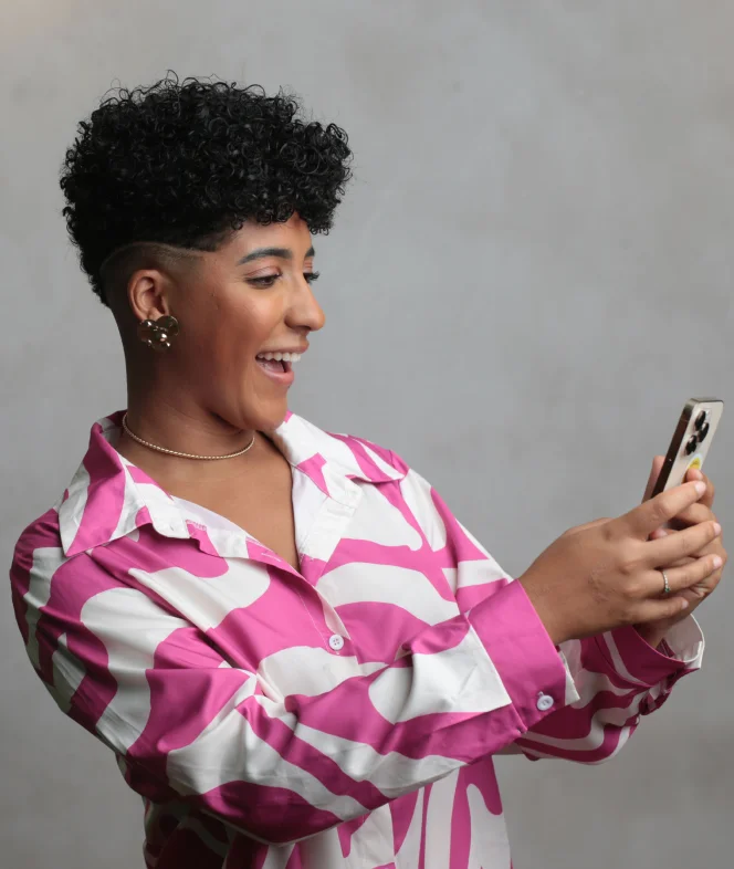 Nath Finanças, mulher negra de cabelo cacheado curto, sorrindo enquanto olha para o celular, vestindo uma camisa rosa e branca estampada.
