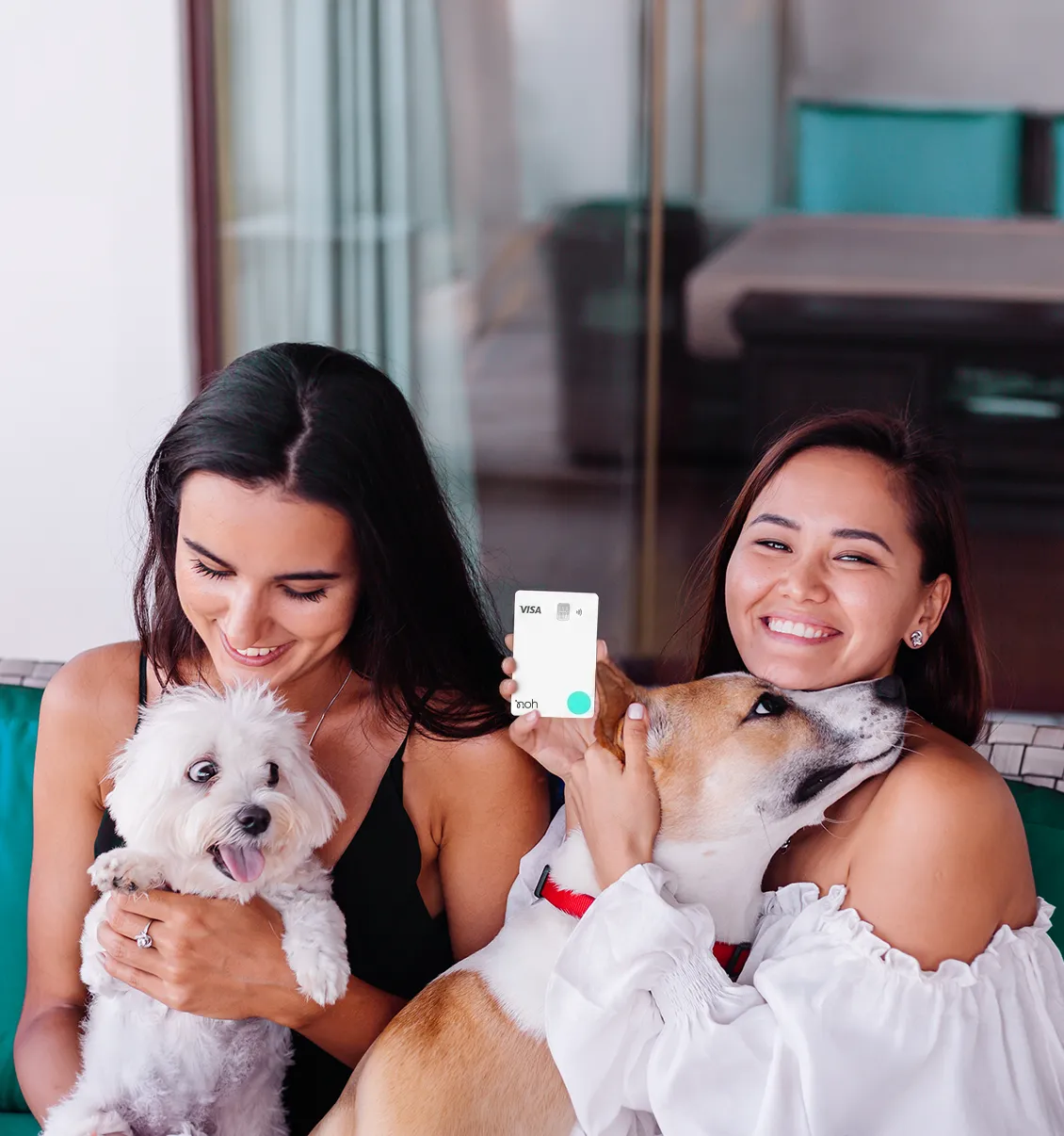 Duas mulheres estão sorrindo sentadas em um sofá, cada uma com um cachorro no colo e uma delas tem um cartão de crédito na mão.
